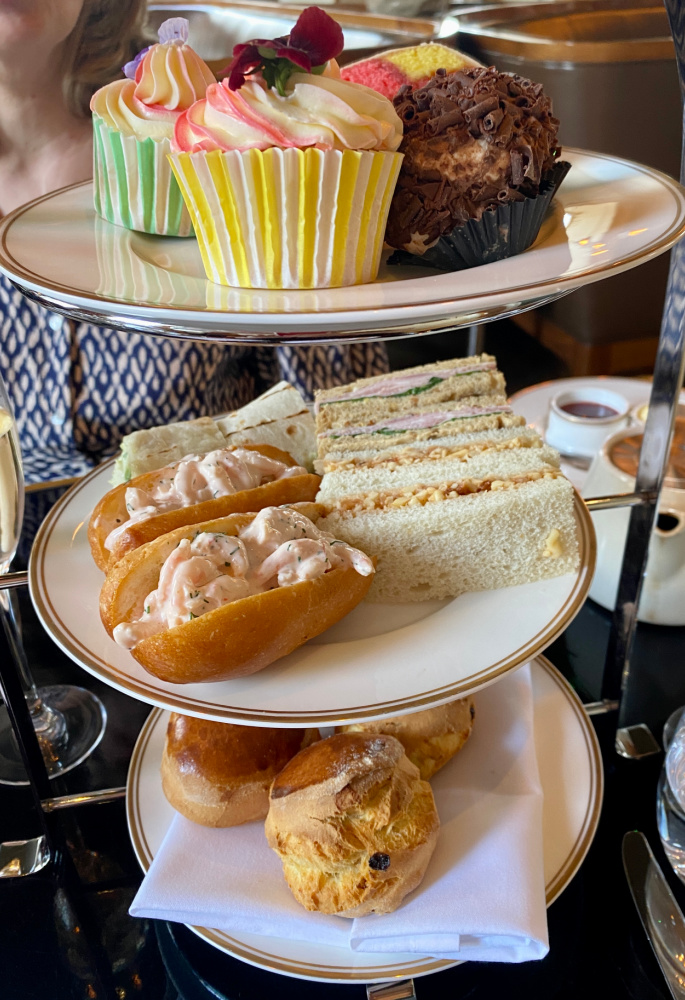 a three tier afternoon tea stand filled with savoury treats, scones and on the top cupcakes and  slices of battenburg cake