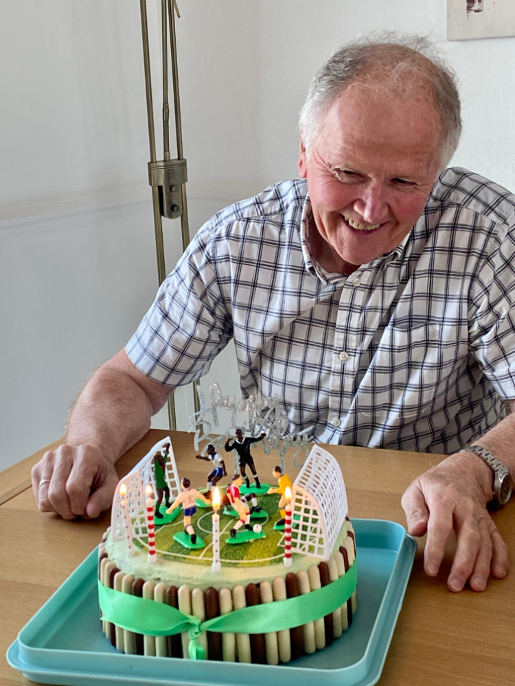 a man in a checked shirt looking down at a round cake topped with a football pitch and some football figurines