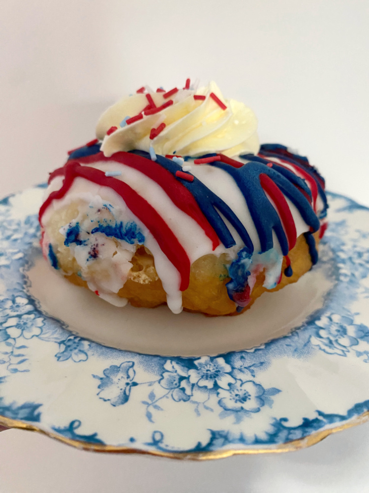 a coronatiion donut drizzled with red white and blue icing and topped off with a blob of cream and red white and blue sprinkles