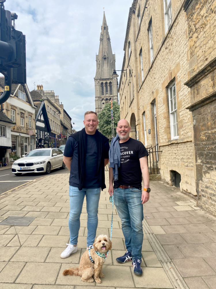 two men standing on a street with a spire in the background. A little dog is sitting in front of them.