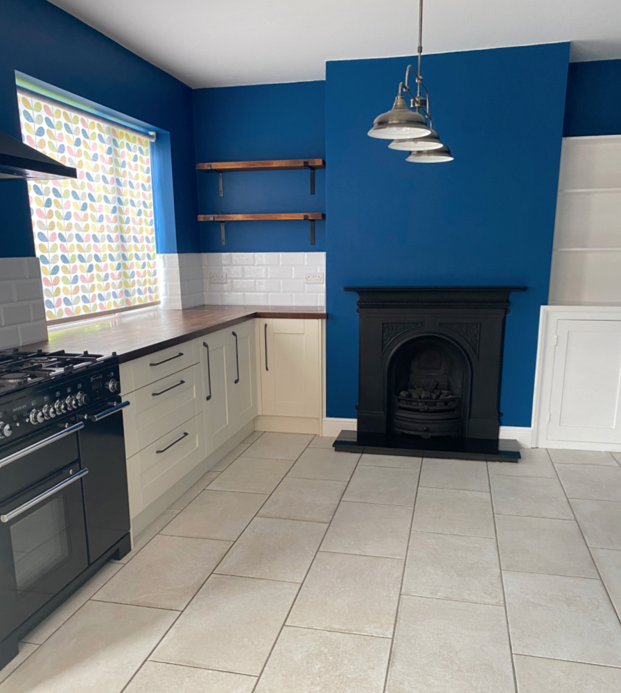 looking into a kitchen with white units on one side, deep blue walls and a victorian fireplace at one end