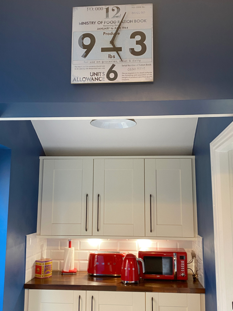 square clock on blue kitchen wall with white cupboards and a wooden shelf with red toaster and kettle in the background