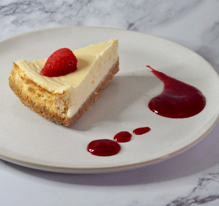 slice of lemon cheesecake on light coloured plate with a splodge of deep red berry coulis