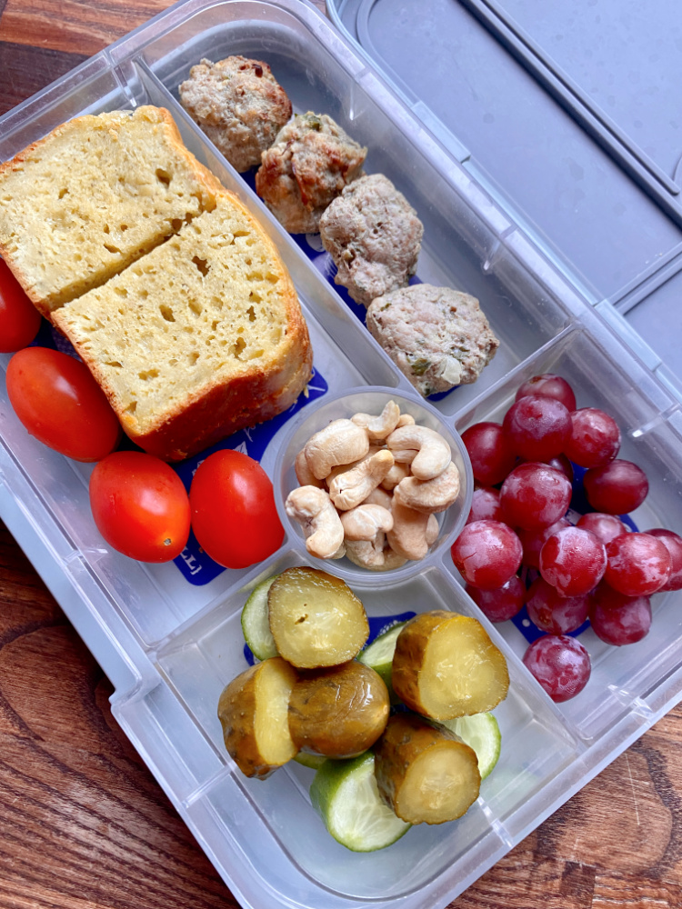 bento lunch box with cashews, grapes, chopped pickles, meatballs and 2 slices of pesto bread
