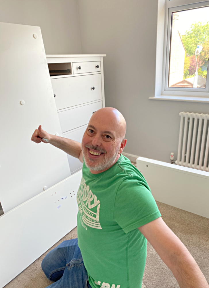 man kneeling among unassembled flat pack furniture. He is smiling and making a thumbs up in the air.