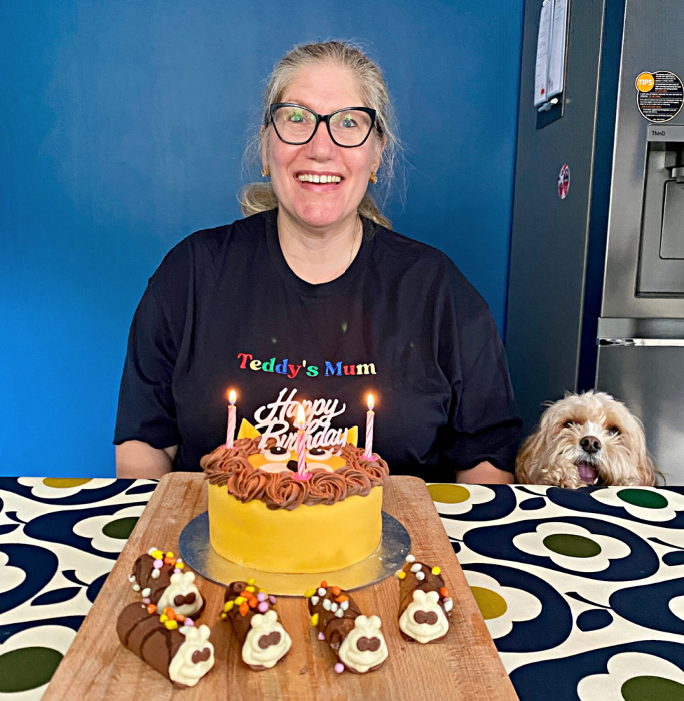 woman sitting behind a lion themed birthday cake. A little dog is next to her trying to get to the table.