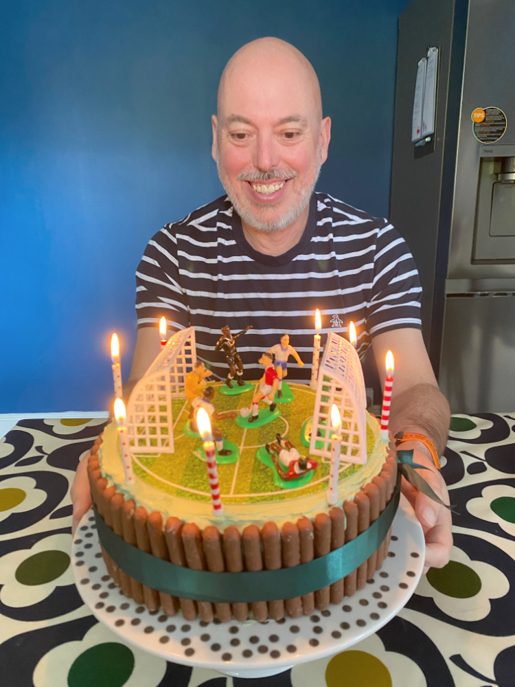 man in stripy tee shirt looking down at candles on his round football pitch themed birthday cake