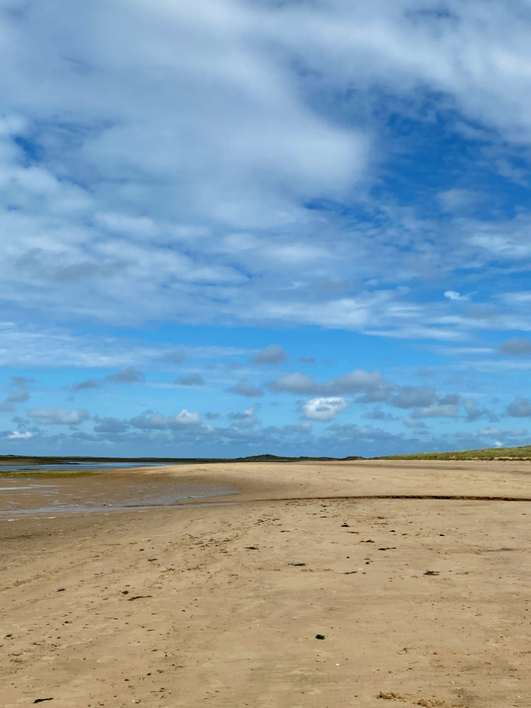 looking along brancaster beach where the sand meets the horizon. There are blue skies and white fluffy clouds in the sky.