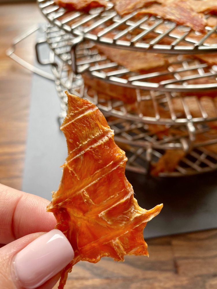 a hand holding a piece of chicken jerky with racks of dehydrated chicken slices in background