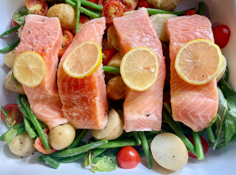 4 uncooked salmon fillets each topped with a slice of lemon lying on a bed of green beans, tomatoes and baby new potatoes