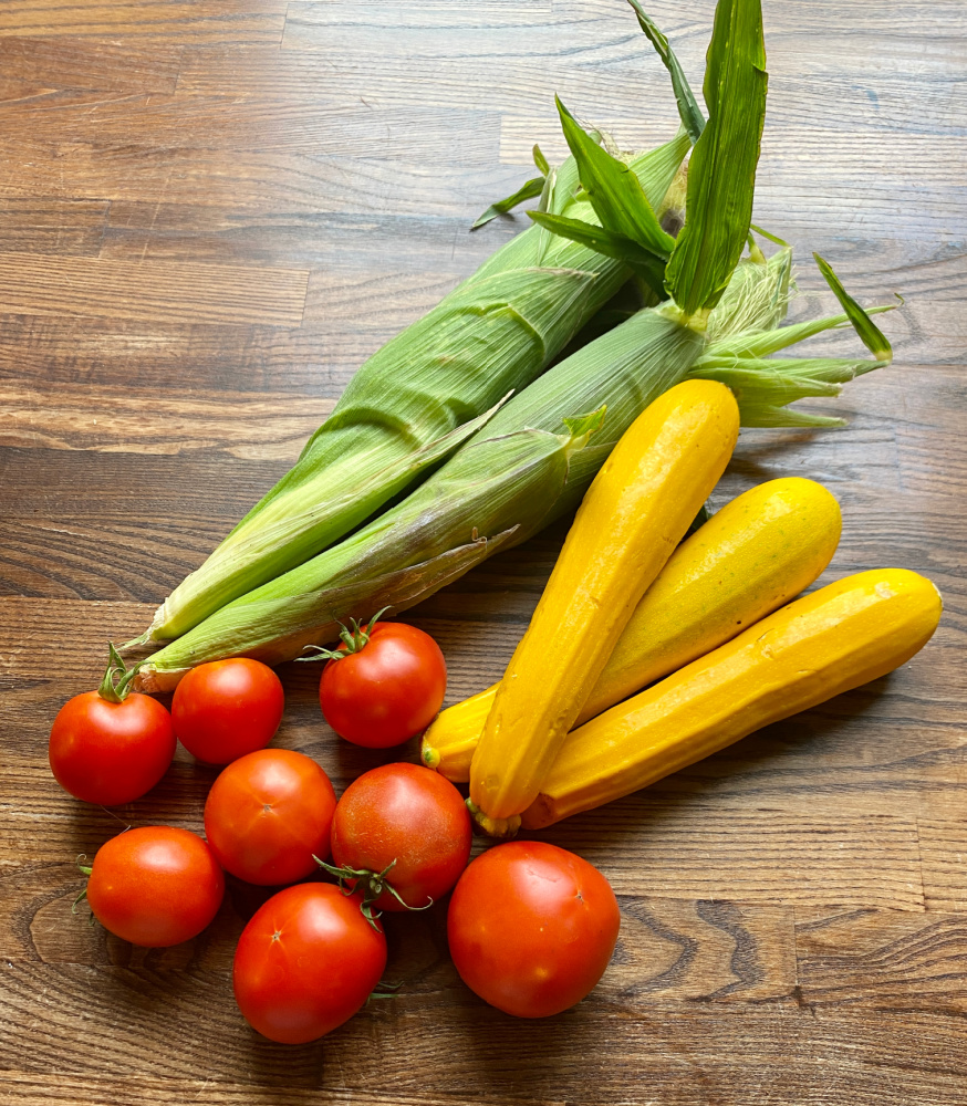 a handful of tomatoes, two ears of corn and three yellow zucchini on a wooden bench