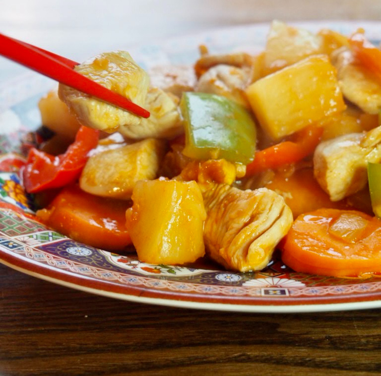 a pair of red chopsticks picking up a piece of chicken from a plate of sweet and sour chicken stir fry with peppers, carrots and pineapple