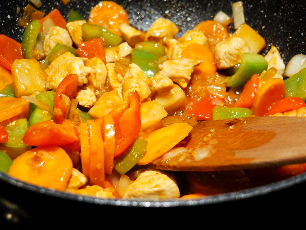 sweet and sour chicken stir fry in a wok with diced chicken, red and green peppers, sliced carrots and pineapple