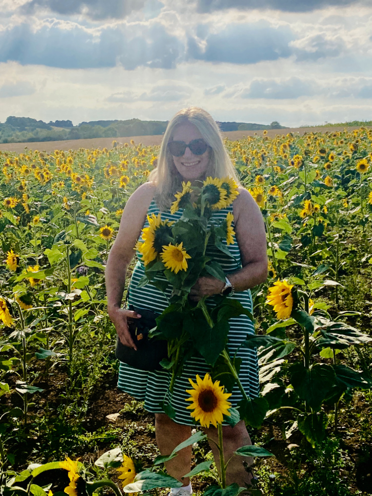 A Woman with long blonde hair wearing a green and white stripe dress holding a bunch of sunflowers in a field of sunflowers