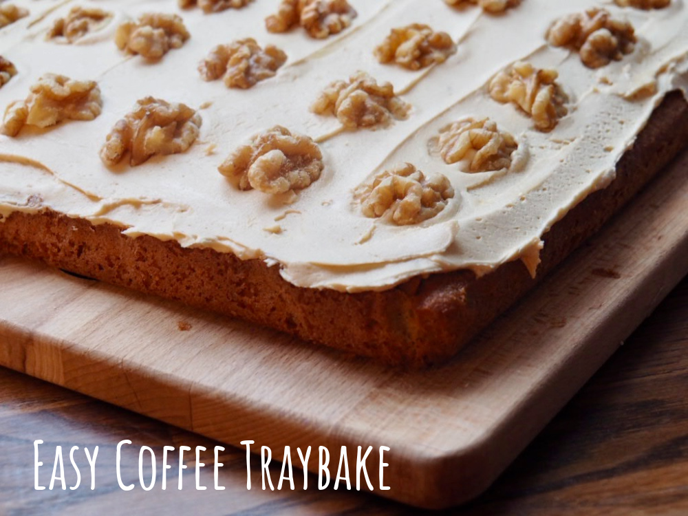 corner of a coffee traybake covered with fudge icing and walnut pieces on a wooden board